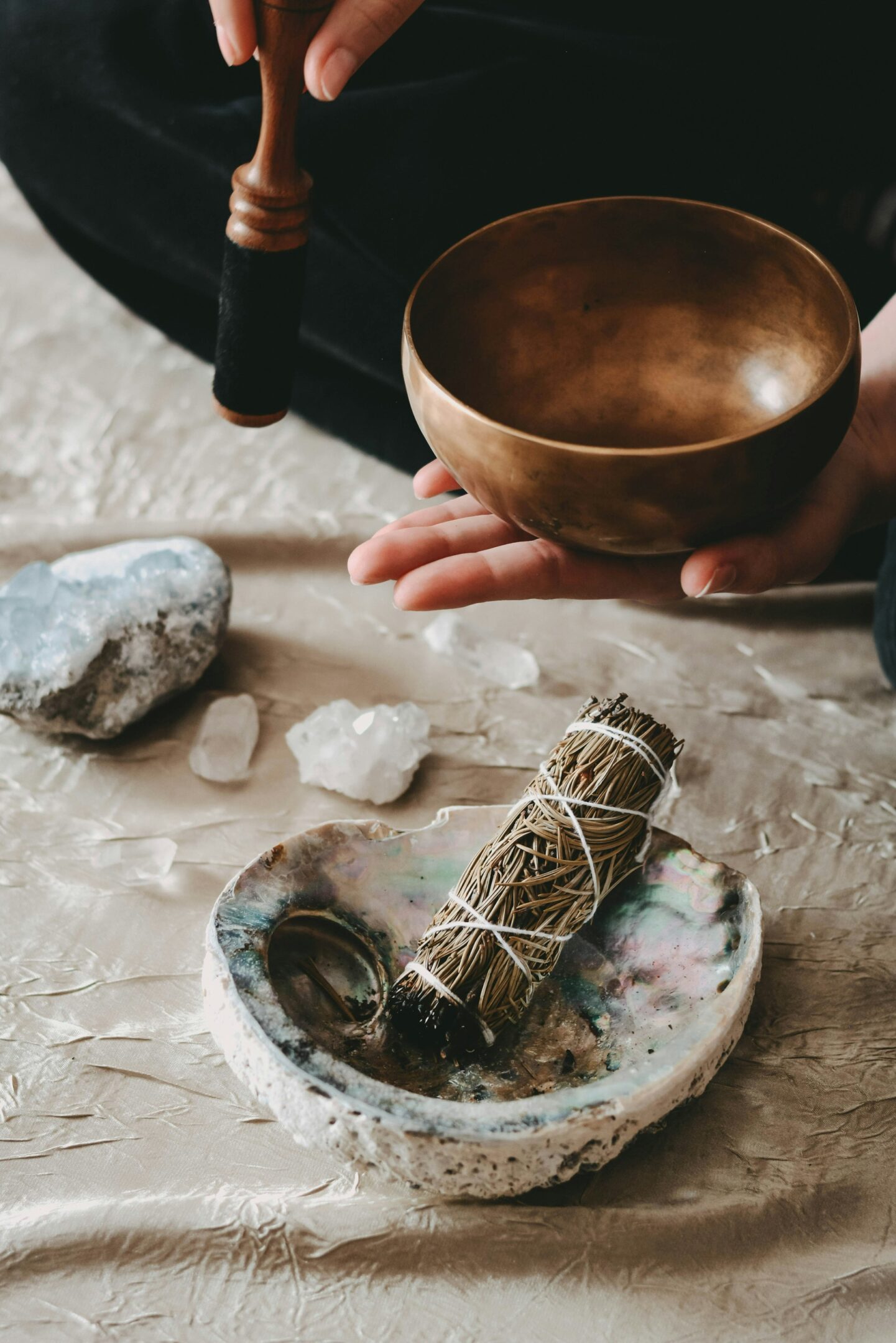 A person holding a singing bowl with crystals and sage on a table.