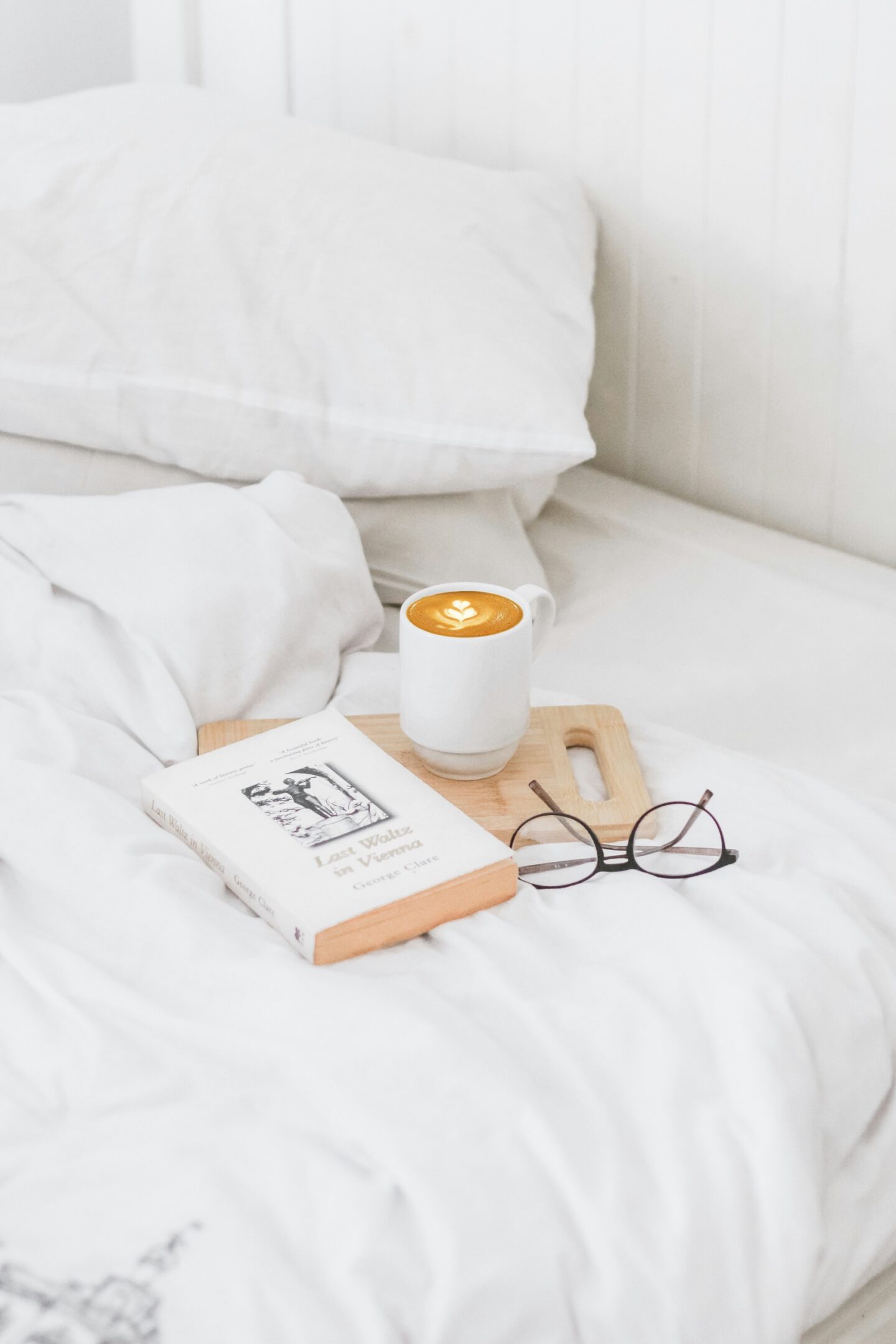 A cozy white bed with a cup of coffee, glasses, and a book.