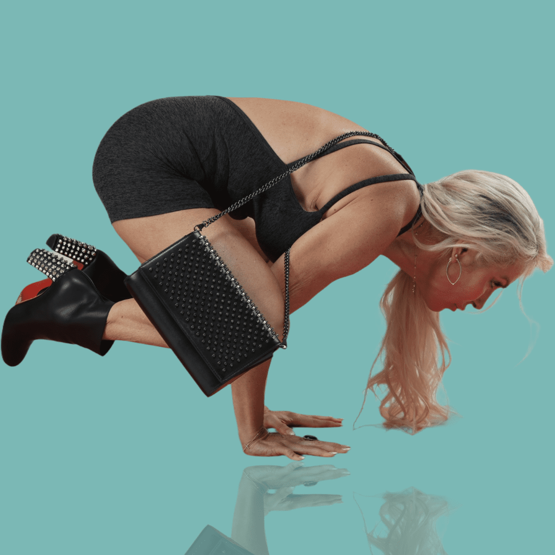 Woman balancing on hands in yoga pose, wearing heels and holding a clutch.